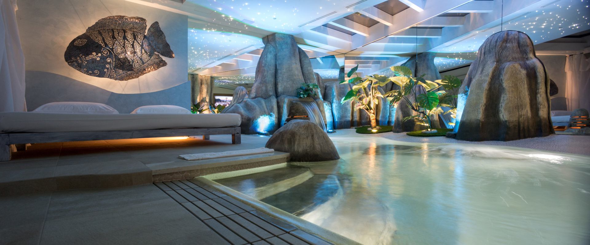Motel K Pool Villa Seychelles vista sulla piscina idromassaggio con cascata e cromoterapia, coreografia di rocce naturali, letto matrimoniale a bordo piscina e piante esotiche, camera a tema isole dell'oceano indiano